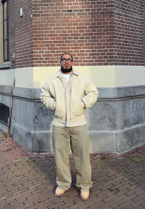 Uomo con occhiali, giacca beige chiaro, camicia a righe, pantaloni larghi e stivali, in piedi con le mani in tasca all'angolo di un edificio in mattoni e pietra.