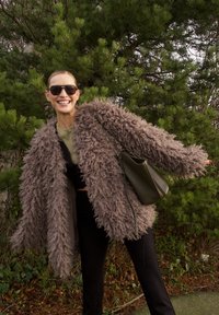 Manteau texturé, marron et duveteux avec manches longues, porté sur un haut vert et un pantalon noir ; comprend un sac à bandoulière vert et des lunettes de soleil.