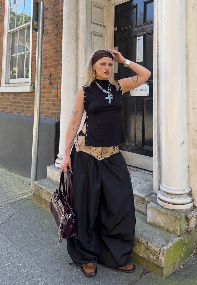 Femme aux cheveux blonds portant un haut noir sans manches, un pantalon noir ample, un foulard et des bijoux, debout près d'une porte noire et des marches en pierre.