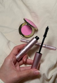 Too Faced mascara in een metallic roze tube, zwarte borstel blootgesteld; roze blushcompact met gouden rand en verhoogde tekst op de achtergrond.