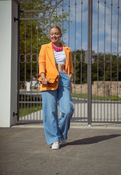 Blazer naranja brillante sobre una blusa corta blanca, combinado con jeans de pierna ancha azul claro. Lleva un clutch naranja y presenta un collar texturizado.