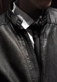 Giacca in pelle nera con un distintivo colletto a costine, abbinata a una camicia bianca e una cravatta nera. Primo piano che mostra la texture della giacca e la cerniera.