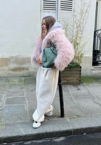 Une femme se tient sur un trottoir en ville, portant une veste rose duveteuse, un pantalon blanc large, des baskets blanches, et tenant un sac à main vert.