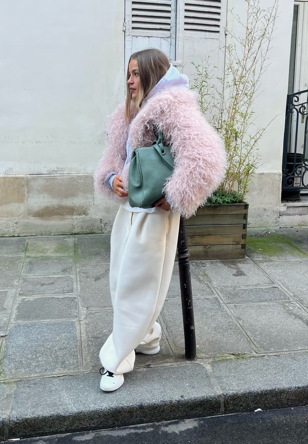 Kvinde står på en byfortov iført en pink fluffy jakke, hvide vide bukser, hvide sneakers og bærer en grøn håndtaske.