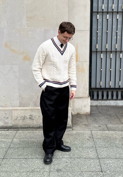 Suéter de punto trenzado en color crema, con escote en V y detalles a rayas. Combinado con pantalones de pierna ancha negros y zapatos negros.