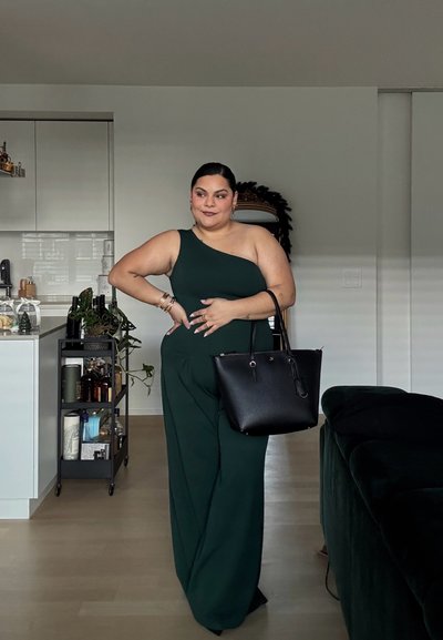 Jumpsuit verde de un solo hombro con piernas anchas. Bolsa negra con acentos dorados. Decoración minimalista en el fondo. Luz natural.