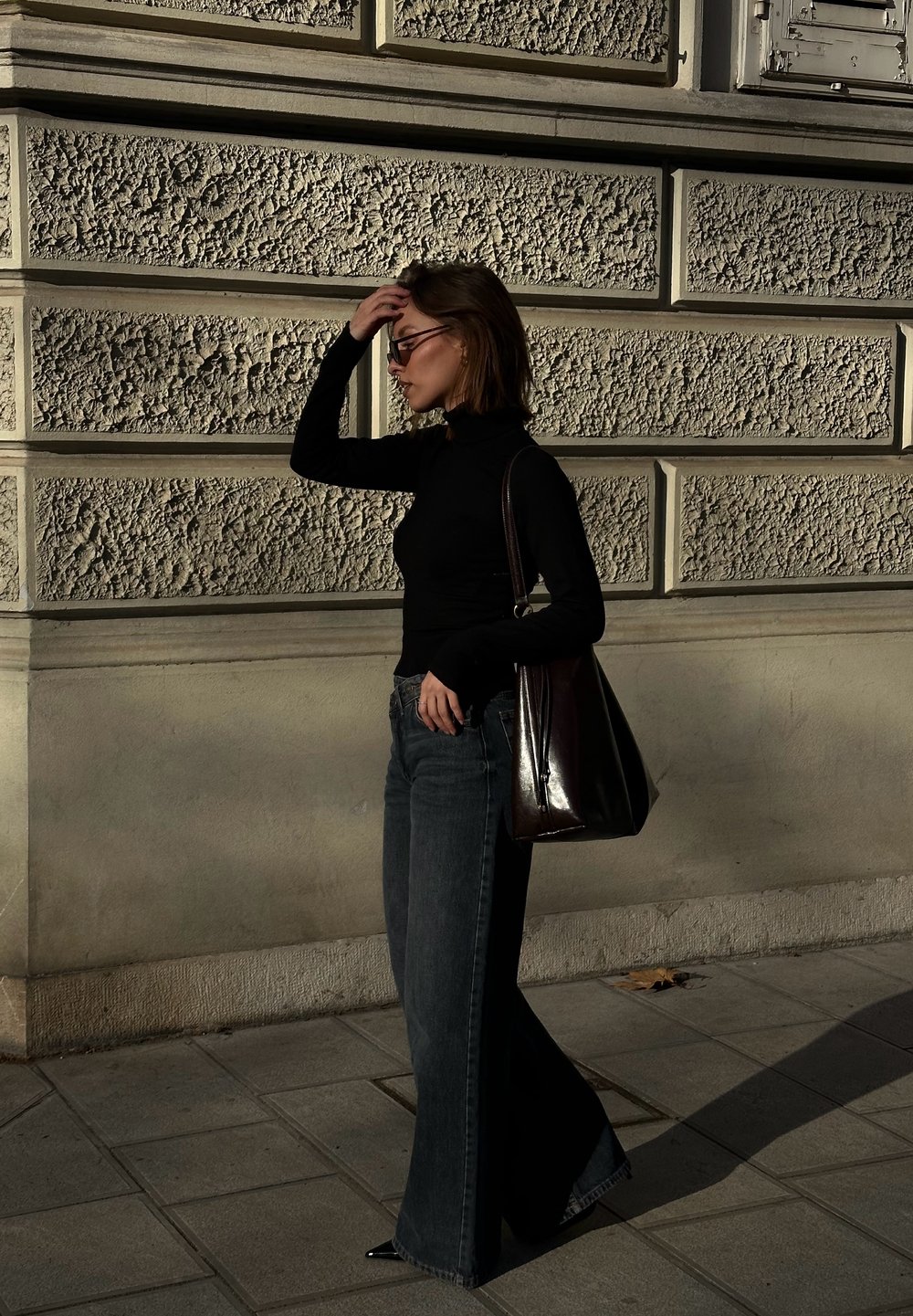 Donna con dolcevita nero e jeans a gamba larga, che porta una borsa grande e scura, camminando e toccandosi i capelli accanto a un muro di pietra con texture.