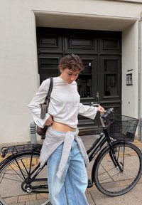 Balta krekla ar garām piedurknēm un kabatu, ar zvana formas piedurknēm, saskaņota ar gaismaziliem plaša griezuma biksēm un pelēku džemperi, kas sasiets ap vidukli, blakus melnai velosipēdam.