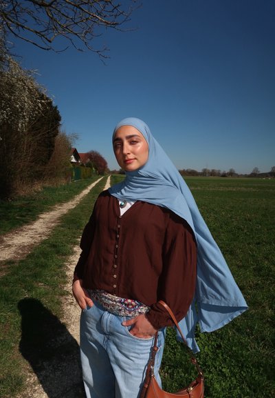 Camisa marrón de botones, hijab azul claro, cinturón estampado, jeans azules relajados, bolso marrón; paisaje natural y cielo azul claro de fondo.