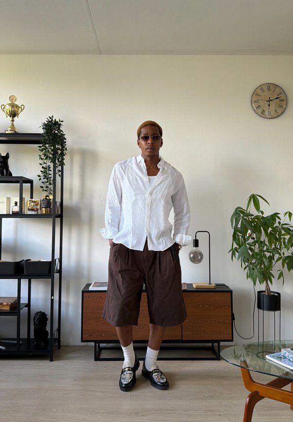 Homme portant des lunettes de soleil, une chemise blanche et un short marron se tient dans une pièce moderne avec des plantes, une horloge et une étagère noire avec des décorations.