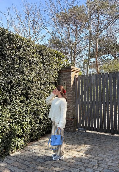 Mujer con suéter blanco y pantalones de rayas está de pie en un camino empedrado cerca de un seto verde y una puerta de madera, sosteniendo un bolso azul.
