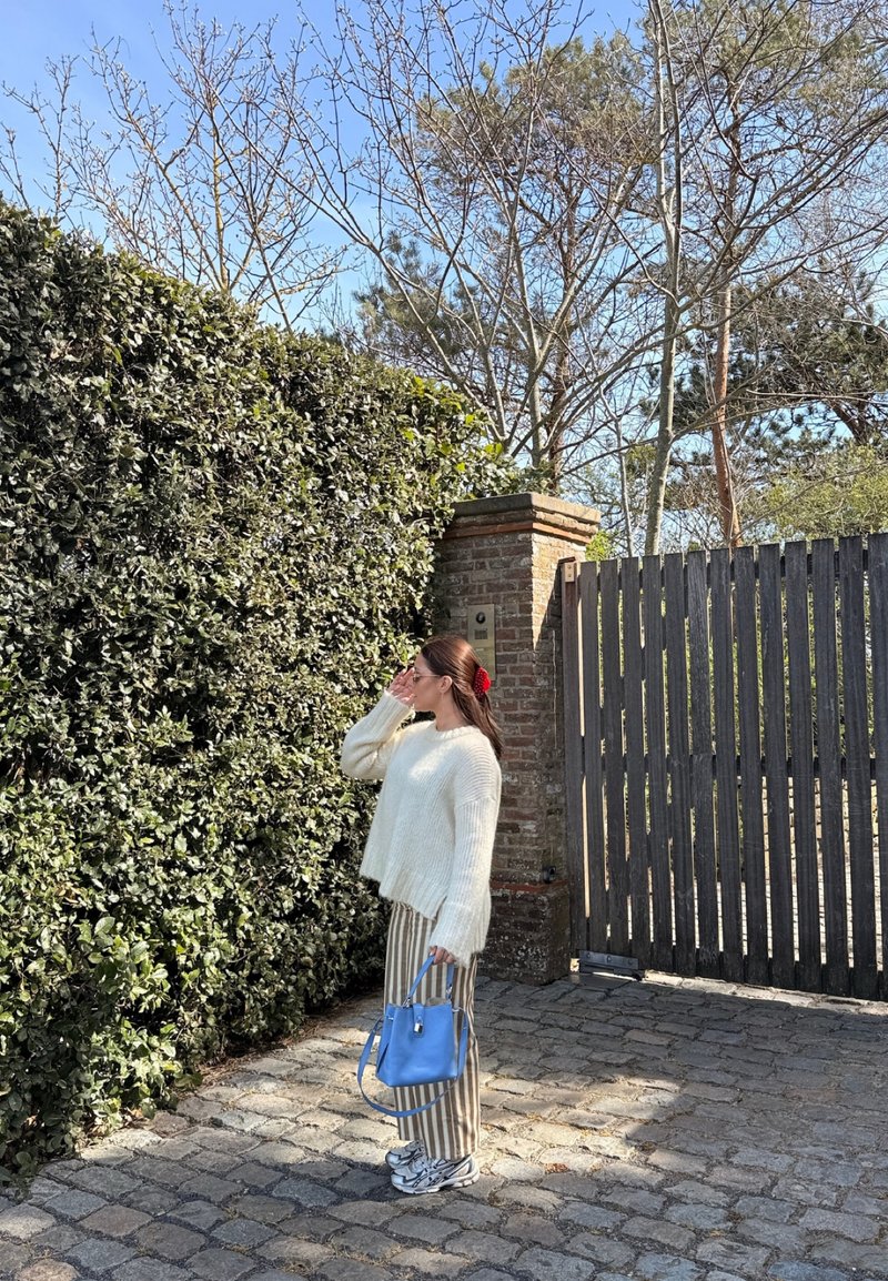 Woman in white sweater and striped pants stands on cobblestone path near green hedge and wooden gate, holding a blue handbag.