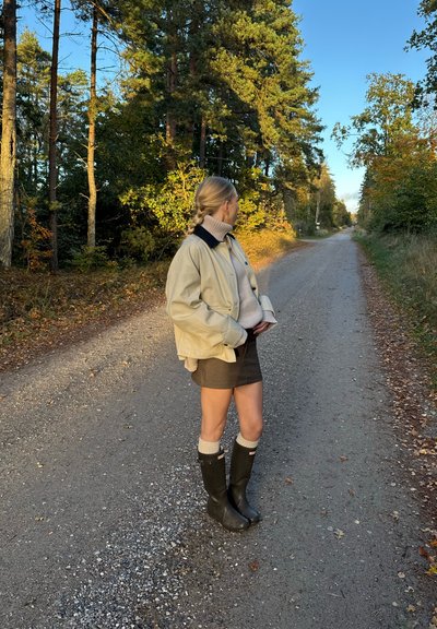 Chaqueta oversized beige claro con un cuello azul marino, suéter de cuello alto gris y mini falda. Botas de goma negras con puños de punto blancos. Fondo de un sendero en el bosque.