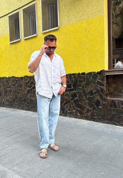 Hombre con una camisa blanca de lino y jeans azul claro, usando gafas de sol y sandalias marrones, de pie en una acera pavimentada gris junto a una pared amarilla.