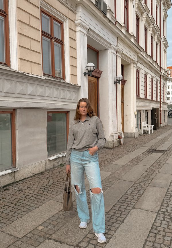 Jeune femme debout sur une rue pavée à côté d'un bâtiment beige, portant un pull gris, un jean clair déchiré, des chaussures blanches et tenant un sac à main marron.
