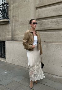 Vrouw in een tan kleurige suede jas, witte top, crèmekleurige rok met kant, tan kleurige laarzen, zwarte zonnebril en met een zwarte handtas op de stoep in de stad.