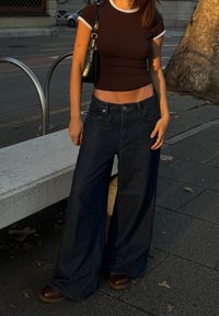 Kvinne i svart crop-top og brede mørke jeans som står på en urban fortau nær en steinbenk og tre.