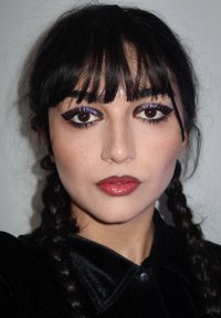 Kobieta z warkoczami i prostą grzywką, nosząca czarną aksamitną koszulę, intensywny eyeliner, fioletowy cień do powiek i błyszczącą czerwoną szminkę.