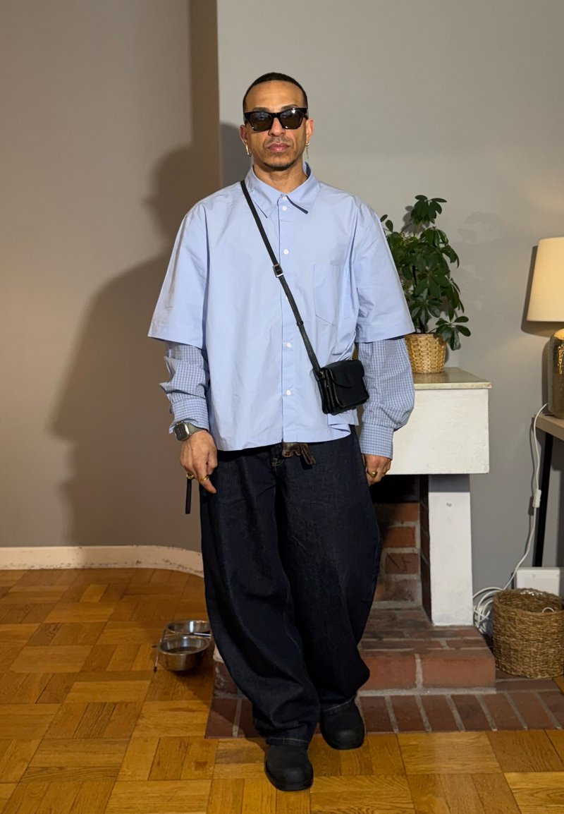 Man wearing layered light blue shirts, dark oversized pants, black sunglasses, crossbody bag, standing indoors near a fireplace and potted plant.