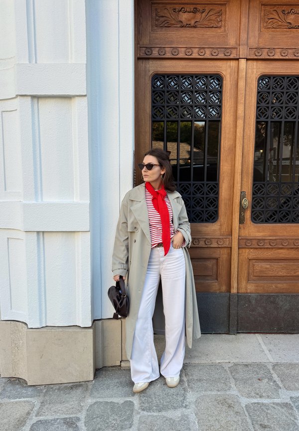 Une femme portant des lunettes de soleil et un trench beige se tient près d’une porte en bois ornée, un sac à main noir à la main. Elle porte une écharpe rouge, un haut rayé et un pantalon blanc.