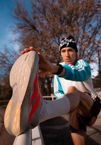 Jeune homme en tenue de sport s'étirant la jambe sur une rambarde métallique en plein air, sous un ciel bleu avec des arbres dénudés en arrière-plan.