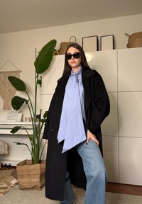 Jeune femme portant un manteau noir, un chemisier bleu, des lunettes de soleil et un jean, debout à l'intérieur près d'une grande plante et de placards blancs.