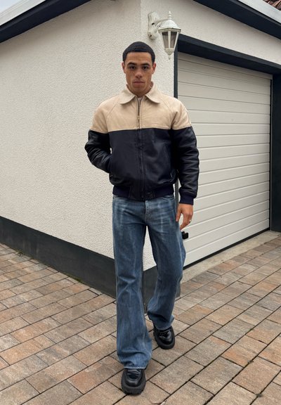 Joven de pie en un camino pavimentado frente a una puerta de garaje blanca, vestido con una chaqueta bicolor, jeans azules y zapatos negros.