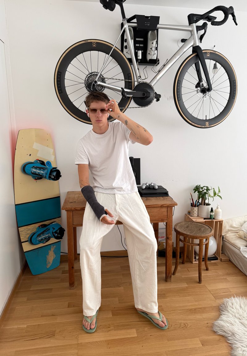 Jeune personne portant des lunettes de soleil et un plâtre au bras gauche, debout dans une pièce avec un vélo accroché au mur et un snowboard appuyé à proximité.