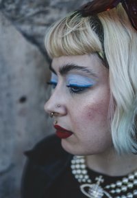Profil av en person med kort, vågigt blont hår, blå eyeliner och rött läppstift, som bär ett pärlhalsband och en septum-piercing.