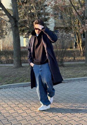 Persona vestida con una sudadera negra bajo un abrigo largo azul marino, pantalones vaqueros holgados azules y zapatillas blancas, caminando por un camino pavimentado a la luz natural.