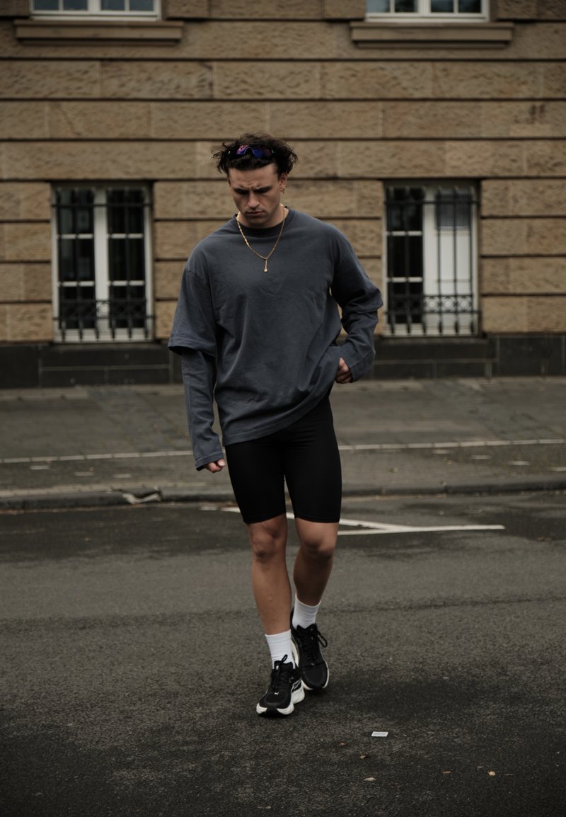 Jeune homme en short noir, chemise oversize gris foncé à manches longues et baskets, marchant sur une rue mouillée avec un bâtiment en arrière-plan.
