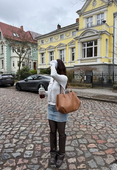 Mujer con suéter blanco y falda de mezclilla sostiene una bebida y se arregla el cabello en una calle empedrada con casas históricas coloridas al fondo.