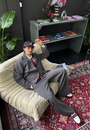 Traje gris de gran tamaño con una corbata burdeos, gorra negra con un logotipo y zapatos blancos. Sentado en un sofá beige con una tela texturizada.