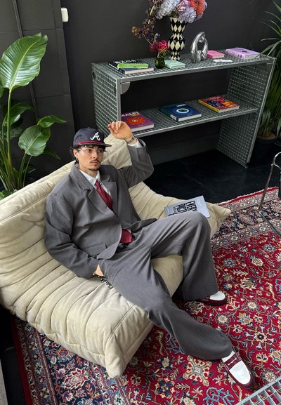 Traje gris de gran tamaño con una corbata burdeos, gorra negra con un logotipo y zapatos blancos. Sentado en un sofá beige con una tela texturizada.