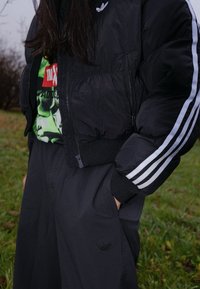 Doudoune noire au design court, accents blancs rayés sur les manches, associée à un t-shirt graphique vert et un pantalon sombre.