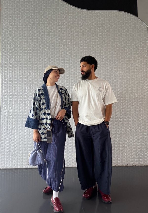 Dvoje ljudi stoje jedno nasuprot drugome, nose ležernu široku odjeću s crvenim cipelama, nasuprot bijelom zidu s heksagonalnim pločicama.