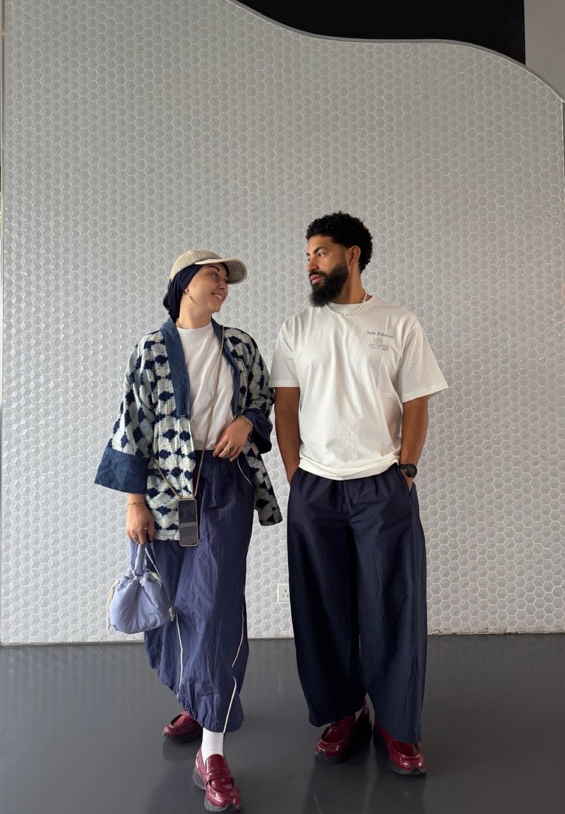 Deux personnes debout face à face, vêtues de vêtements décontractés et amples avec des chaussures rouges, devant un mur blanc carrelé hexagonal.