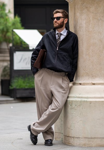 Hombre con gafas de sol, chaqueta negra, corbata y pantalones beige apoyado en una columna de piedra sosteniendo una carpeta de cuero marrón al aire libre.