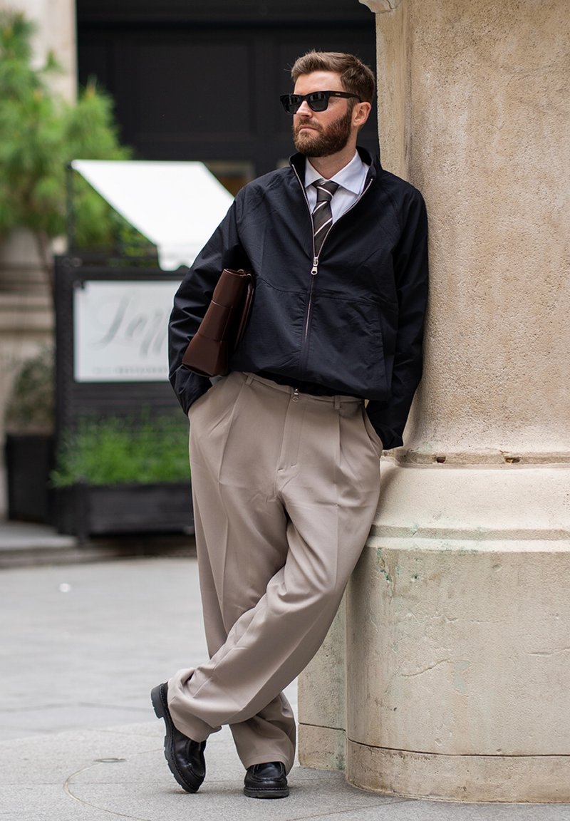 Homme portant des lunettes de soleil, une veste noire, une cravate et un pantalon beige s'appuie contre une colonne en pierre en tenant un portefeuille en cuir marron à l'extérieur.