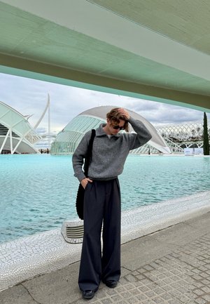 Joven con gafas de sol, suéter gris y pantalones anchos negros, de pie junto a una fuente moderna con arquitectura futurista de fondo.