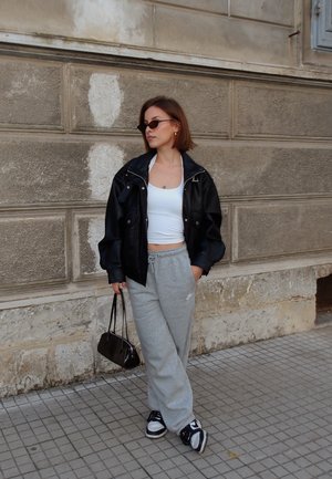 Chaqueta negra de cuero sobredimensionada, top corto blanco, pantalones deportivos grises, bolso negro y zapatillas deportivas en blanco y negro; texturas suaves y estilo casual.