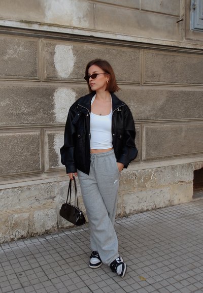 Chaqueta negra de cuero sobredimensionada, top corto blanco, pantalones deportivos grises, bolso negro y zapatillas deportivas en blanco y negro; texturas suaves y estilo casual.
