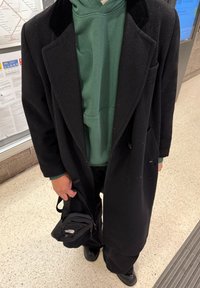 Manteau noir porté sur un sweat à capuche vert, assorti à un pantalon noir. Un petit sac noir est tenu dans une main. Le sol est de couleur claire et texturé.