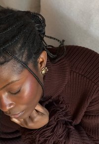 Pull en maille marron avec détails à franges. Boucle d'oreille en manchette dorée. Cheveux foncés tressés. Un éclairage doux met en valeur le ton et la texture de la peau.