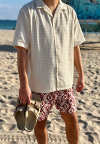 Άτομο που κρατάει μπεζ σανδάλια, φορώντας μπλούζα με υφή σε κρεμ χρώμα με κοντά μανίκια και κόκκινο σορτς με μοτίβα, στέκεται σε αμμώδη παραλία κοντά στον ωκεανό.