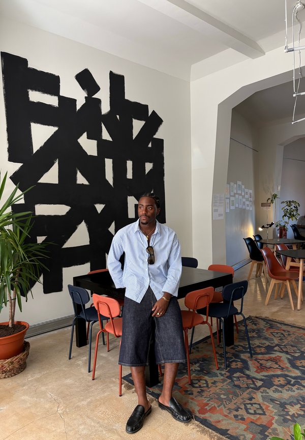 Uomo che indossa una camicia a righe e shorts di jeans seduto su una sedia in un caffè moderno con arte murale astratta nera e piante in vaso.