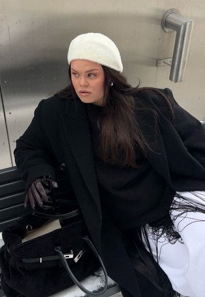 Mujer con boina blanca, abrigo negro y guantes sentada en un banco sosteniendo unas gafas de sol junto a un bolso negro con herrajes plateados.