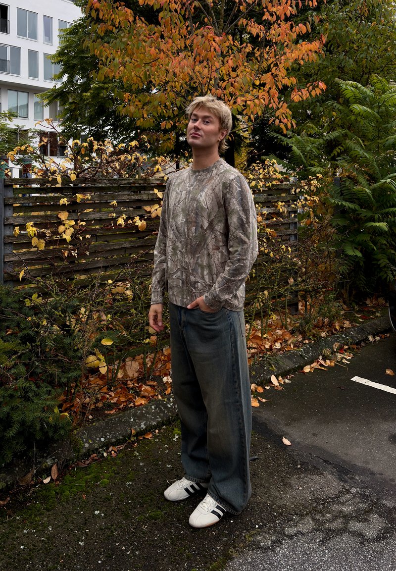 Camouflage lange mouwen shirt en losse blauwe jeans; witte schoenen met zwarte strepen. Omringd door herfstbladeren en houten omheining.