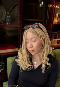Coiffure tressée en blond clair, portant des lunettes de soleil noires sur la tête. Chemise polo noire, assis sur une chaise verte, avec un livre en arrière-plan.