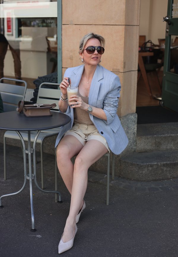 Femeie cu ochelari de soare, sacou albastru deschis și pantaloni scurți bej, stă la o masă în cafenea în aer liber, ținând o cafea cu gheață și poșeta lângă ea.